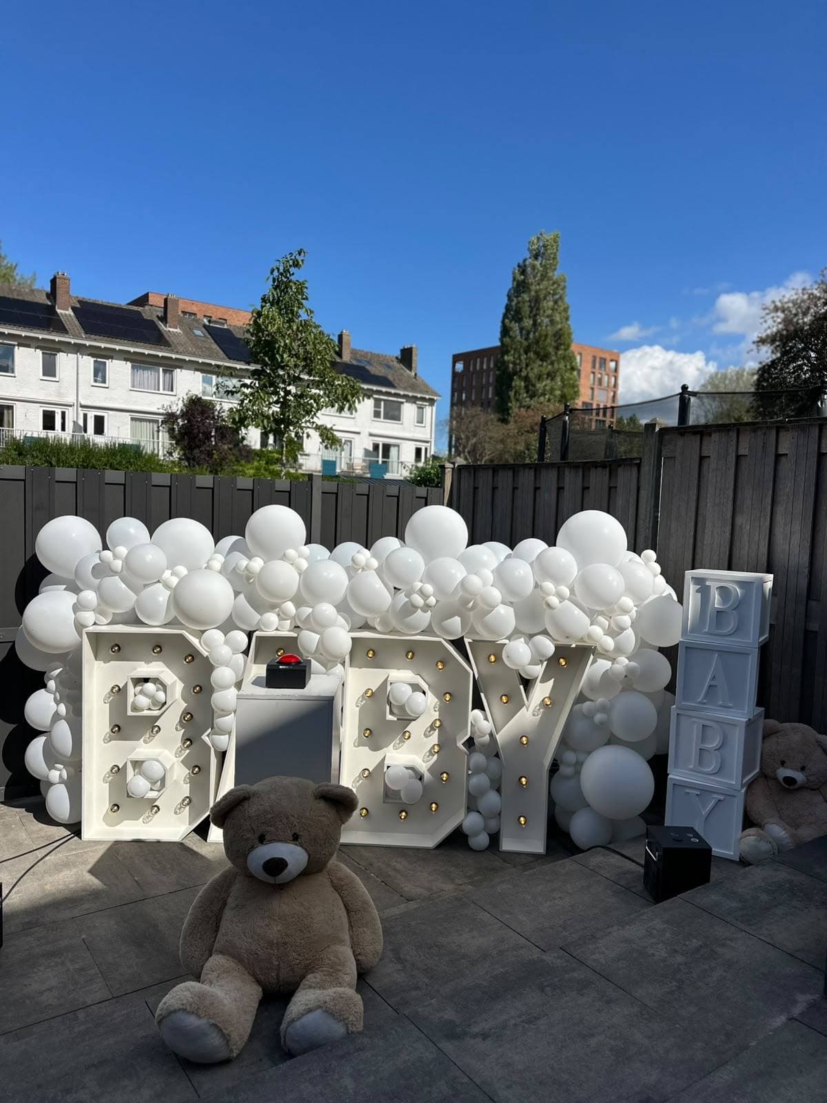 Illuminated BABY marquee letters beneath a cloud of matte white balloons, with stacked white BABY blocks and a teddy bear on a sunny backyard patio between Dutch row houses