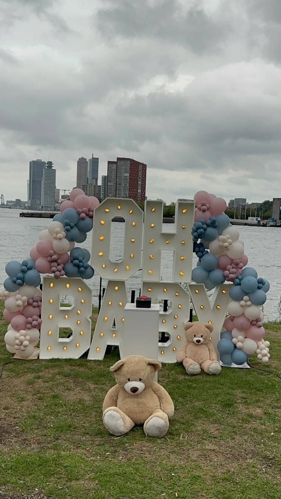 Illuminated OH BABY marquee letters garlanded in dusky pink, dusty blue and ivory balloons, with teddy bears on the riverbank and the Rotterdam skyline across the water