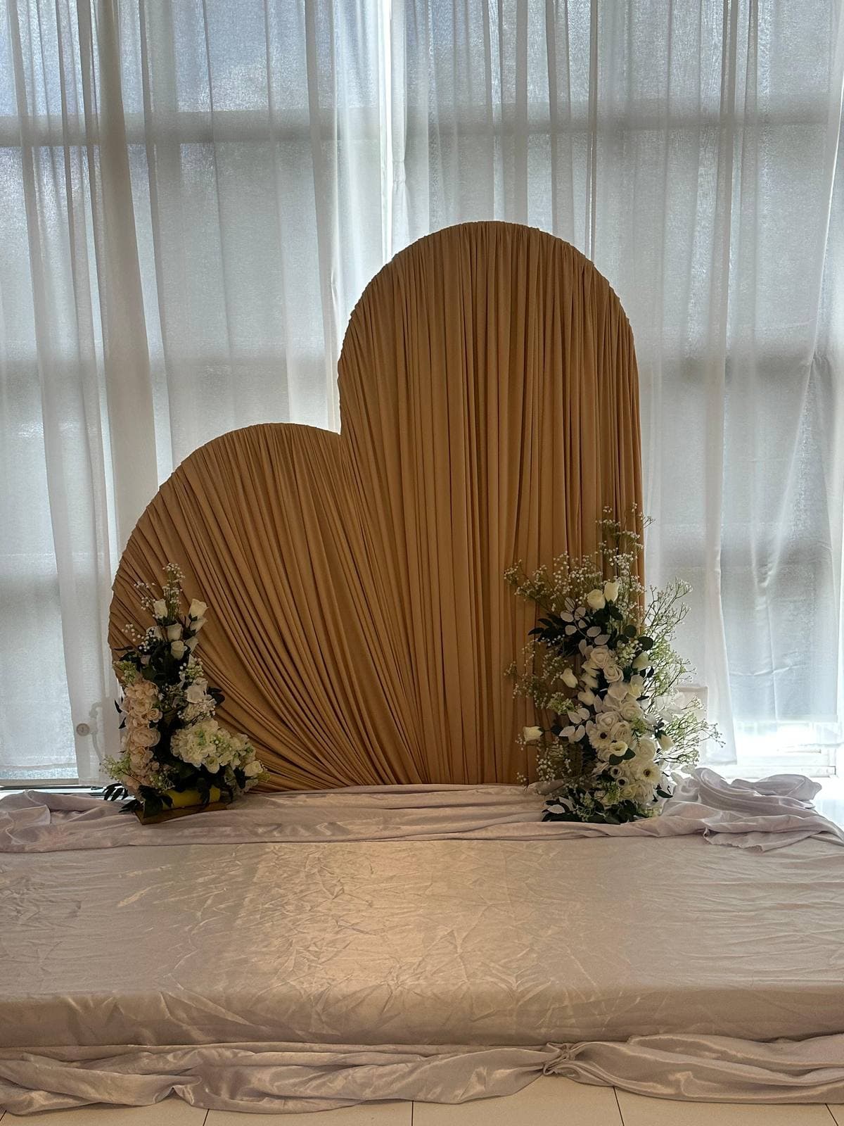 Heart-shaped wedding backdrop of pleated champagne-gold fabric on a white satin-draped plinth, flanked by white rose, hydrangea and baby's breath arrangements against sheer curtains